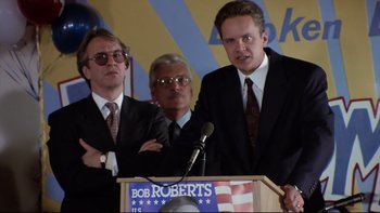 Movie still from “Bob Roberts” (1992), directed by Tim Robbins – A group of men standing at a podium in front of an american flag; Medium shot, Low angle
