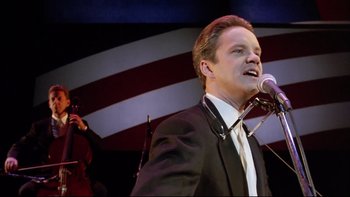 Movie still from “Bob Roberts” (1992), directed by Tim Robbins – A man in a suit and tie holding a microphone; Close Up shot, Low angle