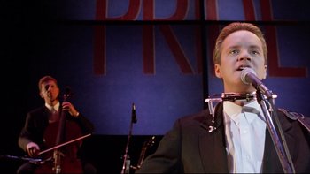 Movie still from “Bob Roberts” (1992), directed by Tim Robbins – A man in a black suit is holding a microphone; Medium shot, Low angle