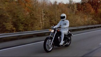 Movie still from “Bob Roberts” (1992), directed by Tim Robbins – A person riding a motorcycle on a road near a forest; Wide shot, High angle