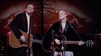 Movie still from “Bob Roberts” (1992), directed by Tim Robbins – A man and a woman are playing guitar and singing; Medium shot, Low angle