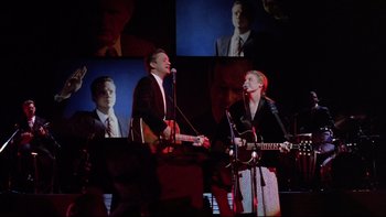 Movie still from “Bob Roberts” (1992), directed by Tim Robbins – A man and a woman singing on stage; Medium shot, Low angle