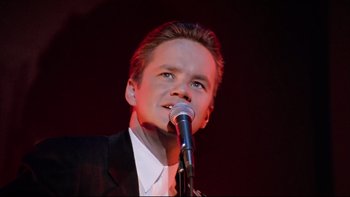 Movie still from “Bob Roberts” (1992), directed by Tim Robbins – A man in a black suit is singing into a microphone; Close Up shot, Low angle