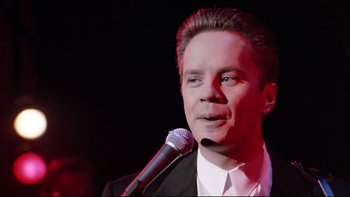 Movie still from “Bob Roberts” (1992), directed by Tim Robbins – A man in a suit and tie holding a microphone; Close Up shot, Low angle