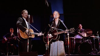 Movie still from “Bob Roberts” (1992), directed by Tim Robbins – A man and a woman singing on stage with guitars; Medium shot, Low angle