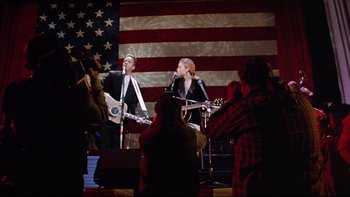 Movie still from “Bob Roberts” (1992), directed by Tim Robbins – Two people are singing and playing guitars in front of an american flag; Medium shot, Low angle