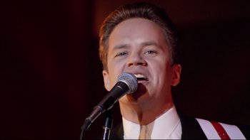 Movie still from “Bob Roberts” (1992), directed by Tim Robbins – A man in a white shirt is singing into a microphone; Close Up shot, Low angle