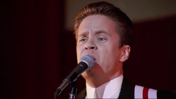 Movie still from “Bob Roberts” (1992), directed by Tim Robbins – A man in a white shirt is singing into a microphone; Close Up shot, Low angle