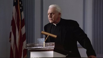 Movie still from “Bob Roberts” (1992), directed by Tim Robbins – A man in a priest's outfit is speaking at a podium; Medium shot, Low angle