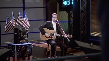 Movie still from “Bob Roberts” (1992), directed by Tim Robbins – A man sitting on a chair holding a microphone and playing a guitar; Medium shot, High angle