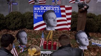 Movie still from “Bob Roberts” (1992), directed by Tim Robbins – A group of people gathered around a sign with a picture of bob roberts on it; Medium shot, High angle