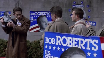 Movie still from “Bob Roberts” (1992), directed by Tim Robbins – A group of men standing next to each other near a building; Medium shot, High angle