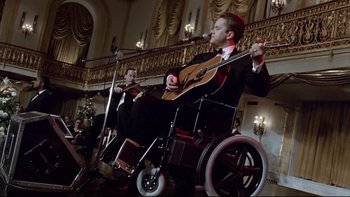 Movie still from “Bob Roberts” (1992), directed by Tim Robbins – A man in a wheel chair playing a guitar and a violin; Wide shot, Low angle