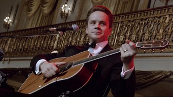 Movie still from “Bob Roberts” (1992), directed by Tim Robbins – A man holding a guitar in front of a microphone; Close Up shot, High angle