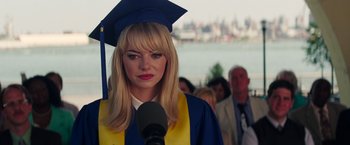 Movie still from “The Amazing Spider-Man 2” (2014), directed by Marc Webb – A woman wearing a graduation cap and gown while speaking into a microphone; Close Up shot, Over the shoulder angle