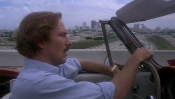 Movie still from “Body Heat” (1981), directed by Lawrence Kasdan – A man driving a car on a highway with a city in the background; Close Up shot, Low angle
