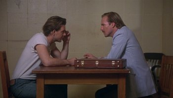 Movie still from “Body Heat” (1981), directed by Lawrence Kasdan – Two men sitting at a table with a briefcase in front of them; Medium shot, Over the shoulder angle