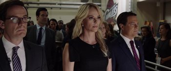 Movie still from “Bombshell” (2019), directed by Jay Roach – A woman in a black dress standing in front of a group of people; Medium shot, Over the shoulder angle