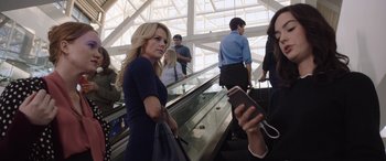 Movie still from “Bombshell” (2019), directed by Jay Roach – A woman is standing on an escalator holding her phone; Medium shot, Over the shoulder angle