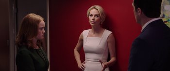 Movie still from “Bombshell” (2019), directed by Jay Roach – A woman in a white dress standing in front of a red wall; Medium shot, Low angle
