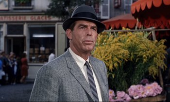 Movie still from “Bon Voyage!” (1962), directed by James Neilson – A man wearing a suit and a hat standing in front of flowers; Medium shot, Over the shoulder angle