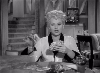 Movie still from “Born Yesterday” (1950), directed by George Cukor – A woman sitting at a table holding a deck of playing cards; Medium shot, Low angle