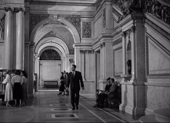 Movie still from “Born Yesterday” (1950), directed by George Cukor – A black and white photo of a man in a suit and tie walking down a hall way; Wide shot, Low angle