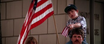 Movie still from “Born on the Fourth of July” (1989), directed by Oliver Stone – A young boy holding a flag in front of an american flag; Medium shot, Low angle