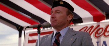 Movie still from “Born on the Fourth of July” (1989), directed by Oliver Stone – A man wearing a hat and a suit speaking into a microphone in front of an american flag; Close Up shot, Low angle