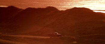 Movie still from “Born on the Fourth of July” (1989), directed by Oliver Stone – A truck driving down a road near a body of water at sunset; Extreme Wide shot, Low angle