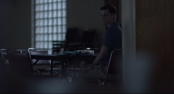 Movie still from “Boy Erased” (2018), directed by Joel Edgerton – A man sitting at a table in a dark room; Medium shot, Over the shoulder angle