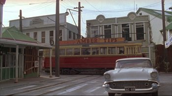 Movie still from “Dead Alive” (1992), directed by Peter Jackson – An old car drives down the street in front of an orange and yellow train; Extreme Wide shot, Low angle