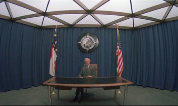 Movie still from “Brainstorm” (1983), directed by Douglas Trumbull – A man sitting at a desk in front of two flags; Wide shot, Low angle