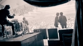 Movie still from “The Beatles: Eight Days a Week - The Touring Years” (2016), directed by Ron Howard – Two men standing in front of an audience in front of a stage; Extreme Wide shot, High angle