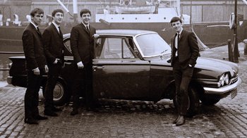 Movie still from “The Beatles: Eight Days a Week - The Touring Years” (2016), directed by Ron Howard – Three young men standing in front of an old car; Wide shot, Low angle