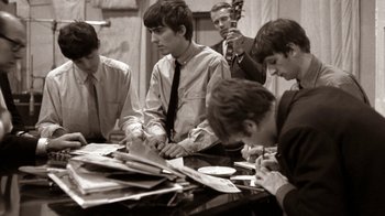 Movie still from “The Beatles: Eight Days a Week - The Touring Years” (2016), directed by Ron Howard – A black and white photo of a group of people sitting at a table; Medium shot, High angle