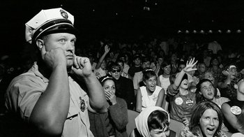 Movie still from “The Beatles: Eight Days a Week - The Touring Years” (2016), directed by Ron Howard – A crowd of people sitting and standing in a room; Medium shot, High angle