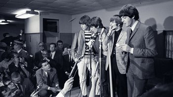 Movie still from “The Beatles: Eight Days a Week - The Touring Years” (2016), directed by Ron Howard – A group of people that are standing in front of a microphone; Wide shot, Over the shoulder angle