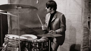 Movie still from “The Beatles: Eight Days a Week - The Touring Years” (2016), directed by Ron Howard – A man wearing a suit and tie playing drums; Medium shot, Low angle