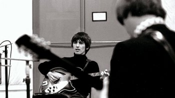 Movie still from “The Beatles: Eight Days a Week - The Touring Years” (2016), directed by Ron Howard – A man playing an electric guitar while another man watches; Medium shot, Over the shoulder angle