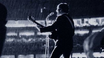Movie still from “The Beatles: Eight Days a Week - The Touring Years” (2016), directed by Ron Howard – A man in a black suit is playing a guitar; Medium shot, Low angle