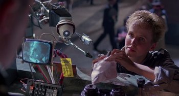 Movie still from “Brazil” (1985), directed by Terry Gilliam – A person sitting at a table with a bag of food; Close Up shot, Over the shoulder angle