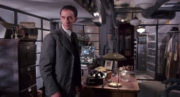 Movie still from “Brazil” (1985), directed by Terry Gilliam – A man in a suit standing in front of a desk; Medium shot, Low angle