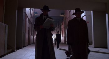 Movie still from “Brazil” (1985), directed by Terry Gilliam – A man in a trench coat and a hat standing in a hallway; Wide shot, Low angle