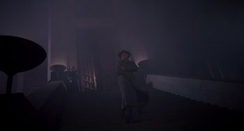 Movie still from “Brazil” (1985), directed by Terry Gilliam – A woman in a hat and a dress standing in a dark room; Wide shot, Low angle