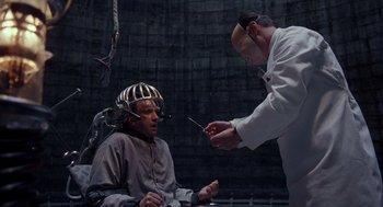 Movie still from “Brazil” (1985), directed by Terry Gilliam – A man wearing a helmet is being examined by a man in a lab coat; Medium shot, High angle