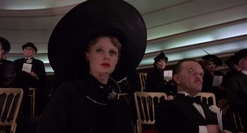 Movie still from “Brazil” (1985), directed by Terry Gilliam – A woman wearing a large black hat in front of two other people; Medium shot, Low angle