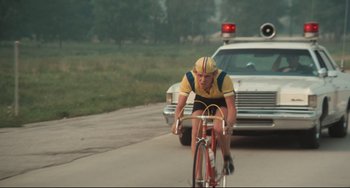 Movie still from “Breaking Away” (1979), directed by Peter Yates – A man riding a bike down a street next to a police car; Wide shot, Low angle