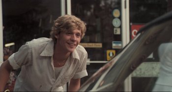 Movie still from “Breaking Away” (1979), directed by Peter Yates – A young man smiling while standing next to a parked car; Medium shot, Over the shoulder angle