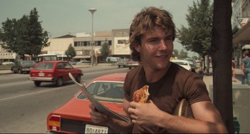 Movie still from “Breaking Away” (1979), directed by Peter Yates – A man holding a piece of pizza while standing in front of a car; Close Up shot, Over the shoulder angle